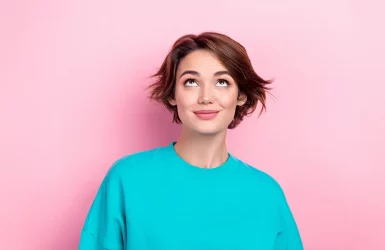 A person with short brown hair in a turquoise shirt looks thoughtfully upward against a pink background.