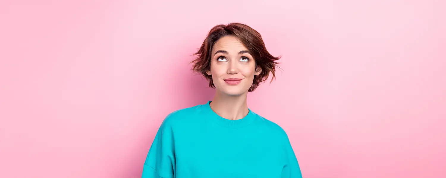 plastic-surgery-hawaii-hero-1 A person with short brown hair in a turquoise shirt looks thoughtfully upward against a pink background.