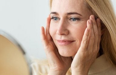 A blonde woman examines her skin in a mirror