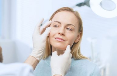 A gloved doctor examines a woman's face under a bright light.