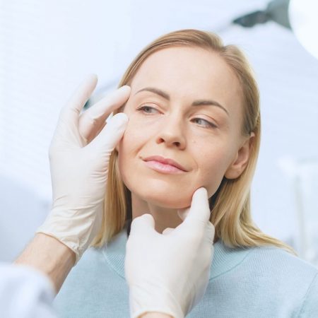 A gloved doctor examines a woman's face under a bright light.