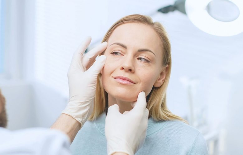 A gloved doctor examines a woman's face under a bright light.