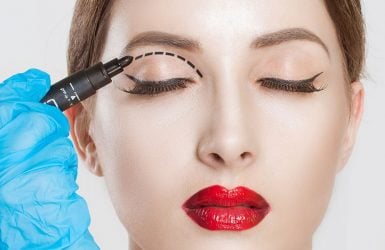 A gloved hand draws a dotted line on a closed eyelid with red lipstick.