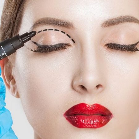 A gloved hand draws a dotted line on a closed eyelid with red lipstick.