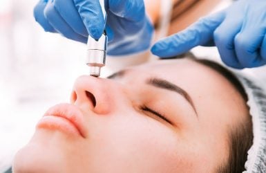 A professional performs dermabrasion on a person's nose in a clinical setting; person wears a hair cover.