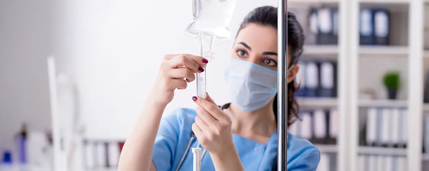 A healthcare worker in blue adjusts an IV drip in a medical facility.