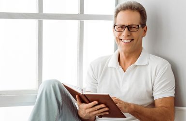 A man in glasses and a white polo smiles by a window