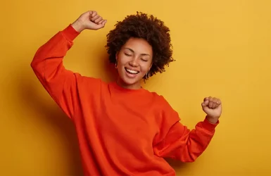 Curly-haired person in orange sweater smiles with eyes closed
