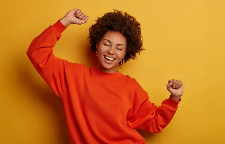 Curly-haired person in orange sweater smiles with eyes closed