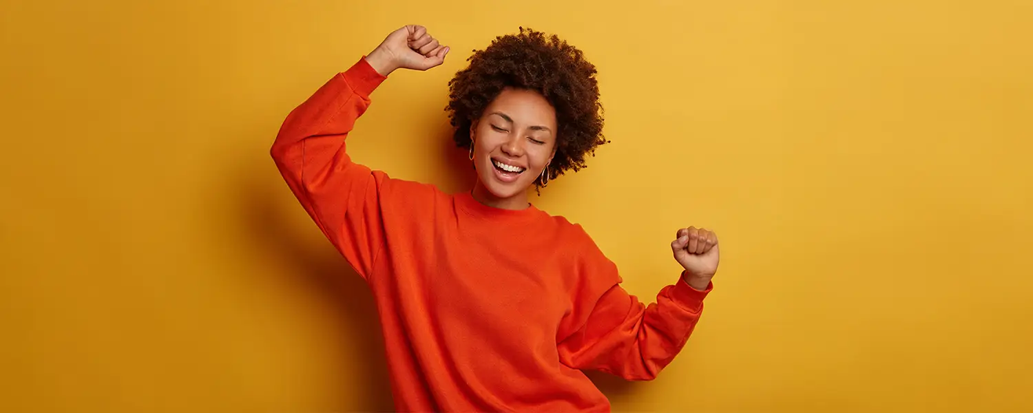 plastic-surgery-hawaii-hero-1 Curly-haired person in orange sweater smiles with eyes closed