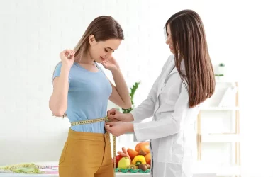 Doctor measures woman's waist; health focus with fruits and vegetables in background.