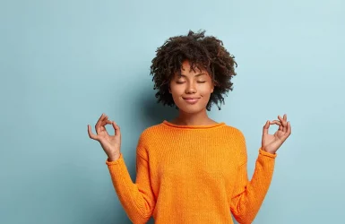 Curly-haired person in orange sweater meditates with closed eyes against a light blue background.