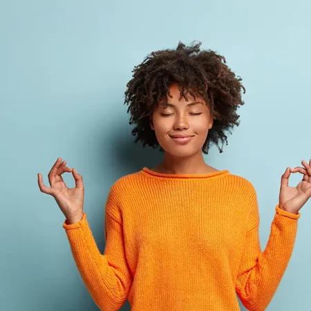 Curly-haired person in orange sweater meditates with closed eyes against a light blue background.