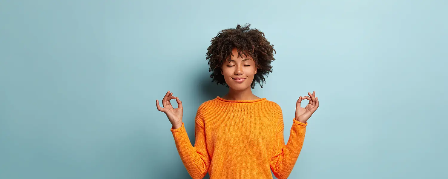 Curly-haired person in orange sweater meditates with closed eyes against a light blue background.