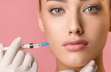 Person with light skin and green eyes receives lip augmentation; syringe held near lips against pink background.