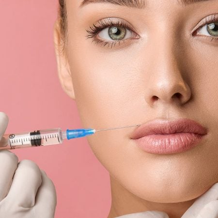 Person with light skin and green eyes receives lip augmentation; syringe held near lips against pink background.