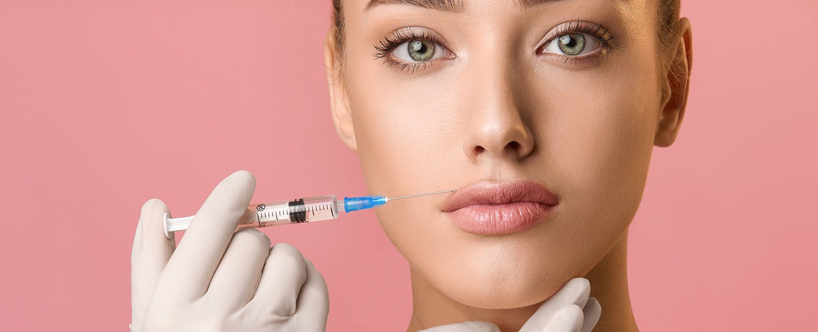 Person with light skin and green eyes receives lip augmentation; syringe held near lips against pink background.