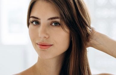 A person with long brown hair and blue eyes touches their hair