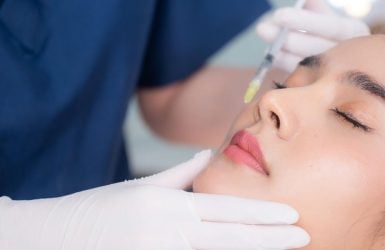 A healthcare professional in scrubs injects a patient's cheek in a clinical setting.