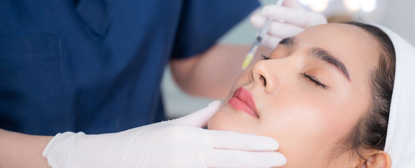 A healthcare professional in scrubs injects a patient's cheek in a clinical setting.