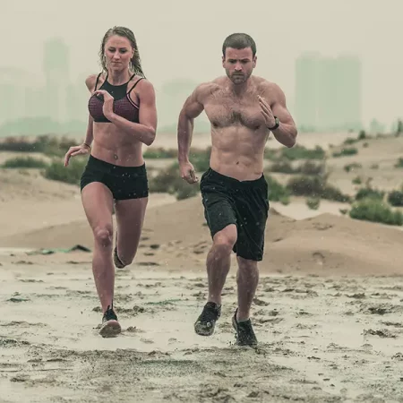 Two people run on a wet beach with a city skyline; overcast sky