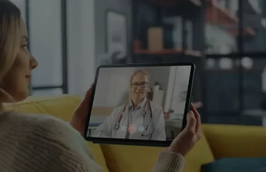 Woman on yellow couch video calls doctor on tablet; modern living room background.