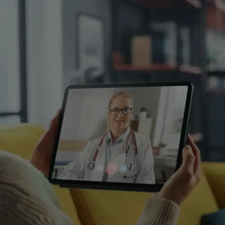 Woman on yellow couch video calls doctor on tablet; modern living room background.