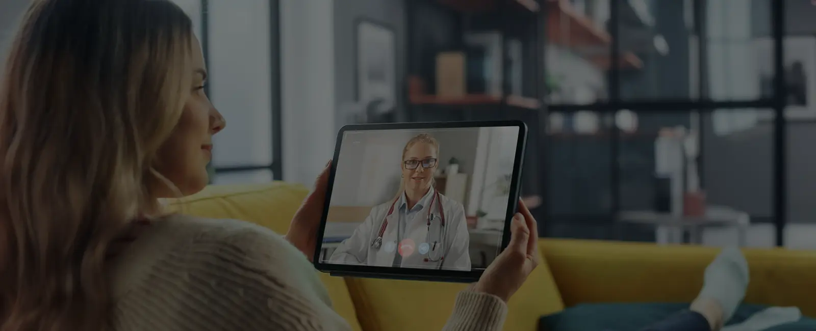 plastic-surgery-hawaii-hero-1 Woman on yellow couch video calls doctor on tablet; modern living room background.
