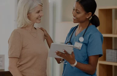 A healthcare worker in blue scrubs talks to an older woman in a well-lit room.