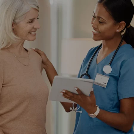 A healthcare worker in blue scrubs talks to an older woman in a well-lit room.